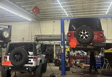 Jeeps being serviced inside 4-Wheel Drive Specialty Co. There is a red Jeep with a black top on a lift with the mechanic working under it and a silver Jeep to the left of it on the concrete. Another vehicle is on a lift in the background slightly cut off from view